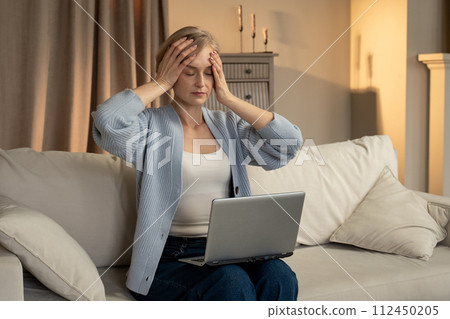 Frustrated Woman Sitting on Sofa With Laptop Covering Her Face With Hand 112450205