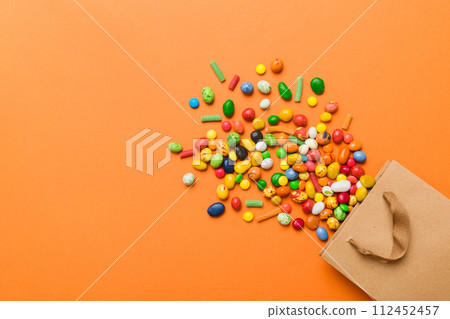 shopping paper gift bag in corner full of assorted traditional candies falling out on colored background with copy space. Happy Holidays sale concept shopping paper gift bag in corner full of assorted traditional candies falling out on colored background with copy space. Happy Holidays sale concept 112452457
