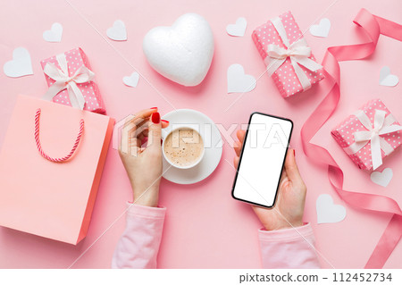 Female hands holding smart phone with coffee for Valentine day, gift box and envelope, hearts Female hands holding smart phone with coffee for Valentine day, gift box and envelope, hearts 112452734