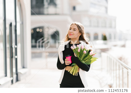 Young woman with a bouquet of spring tulips and a cup of coffee walks down the street in the city 112453176