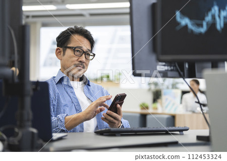 Middle-aged man operating a smartphone in front of a computer 112453324