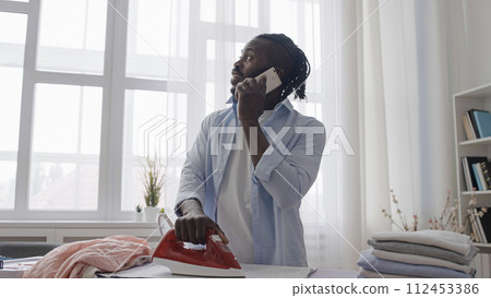 Distracted man talking on phone not paying attention to hot iron on clothes Distracted man talking on phone not paying attention to hot iron on clothes 112453386