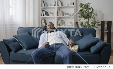 Carefree African American man eating popcorn, lying on sofa and watching TV 112453393