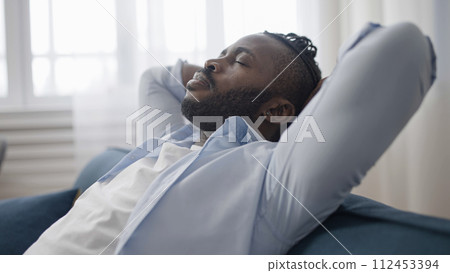 Tired African American man sitting back and relaxing on sofa at home after work day 112453394
