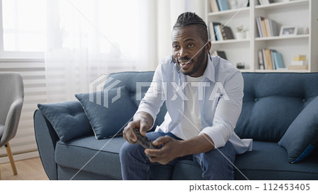 Happy African American man playing video game with a wireless joystick at home Happy African American man playing video game with a wireless joystick at home 112453405