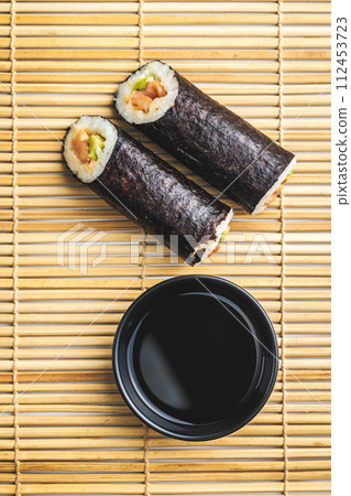 Two Sushi Rolls and a Bowl of soy sauce on a Bamboo Mat 112453723