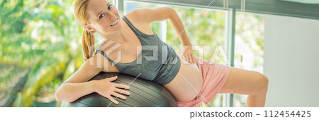 Pregnant woman exercising on fitball at home. Pregnant woman doing relax exercises with a fitness pilates ball. Against the background of the window BANNER, LONG FORMAT 112454425