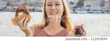 BANNER, LONG FORMAT Woman in Istanbul having breakfast with Simit and a glass of Turkish tea. Glass of Turkish tea and bagel Simit against golden horn bay in Istanbul, Turkey. Turkiye 112454644