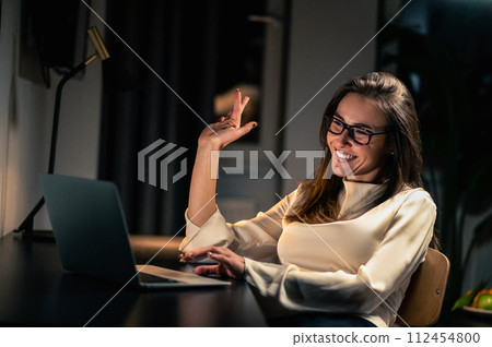 Smiling woman having video call on laptop while sitting at desk at home in evening 112454800
