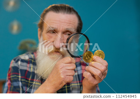 Surprise senior mature man examining golden bitcoins BTC with magnifying glass sitting at table 112454945
