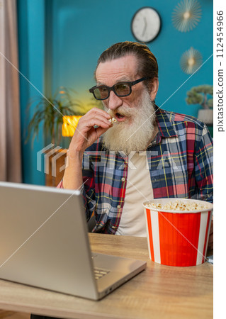 Senior man freelancer taking break from work wearing 3D glasses eating popcorn watching movie film Senior man freelancer taking break from work wearing 3D glasses eating popcorn watching movie film 112454966