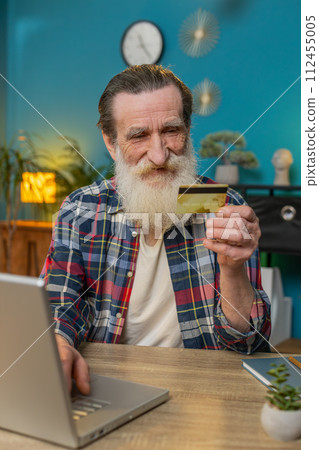 Happy senior man using bank credit card and laptop for online shopping payments at home office Happy senior man using bank credit card and laptop for online shopping payments at home office 112455005