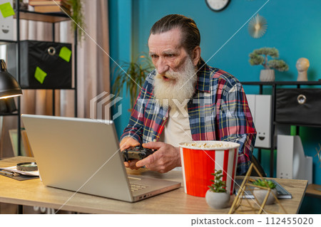 Excited happy senior man celebrating victory in play video game on laptop using controller joystick 112455020