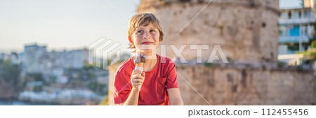 BANNER, LONG FORMAT Boy tourist eating turkish ice cream on background of Hidirlik Tower in Antalya against the backdrop of the Mediterranean bay of the ancient Kaleici district, Turkey. Turkiye 112455456
