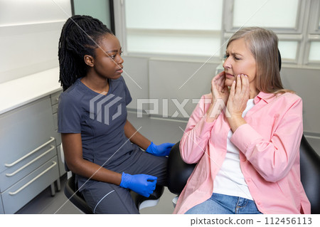 Dentist woman having conversation with blonde middle aged female patient sitting at dental chair Dentist woman having conversation with blonde middle aged female patient sitting at dental chair 112456113