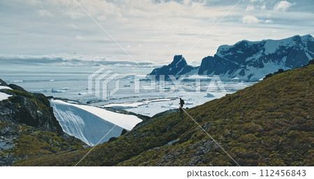 Tourist hiking Antarctica green rock mountain hill down. Ice and snow covered island at hill down. Landscape of polar winter nature at environment preserve scenery. Traveler expedition to South pole 112456843