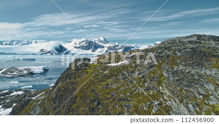Green mountain at Antarctic ocean bay aerial view. Amazing nature of greenery rock at snow covered icebergs background. Birds flying over mount top. Environment scenery of climate change drone shot Green mountain at Antarctic ocean bay aerial view. Amazing nature of greenery rock at snow covered icebergs background. Birds flying over mount top. Environment scenery of climate change drone shot 112456900