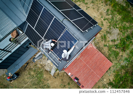 Installers building photovoltaic solar module station on roof of house. Men electricians in helmets installing solar panel system outdoors. Concept of alternative and renewable energy. Aerial view. Installers building photovoltaic solar module station on roof of house. Men electricians in helmets installing solar panel system outdoors. Concept of alternative and renewable energy. Aerial view. 112457846