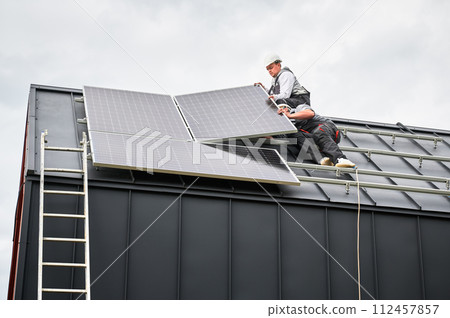 Installers connecting cables while installing photovoltaic solar panels on roof of house. Workers mounting of PV solar modules on rooftop. Concept of alternative and renewable energy. Installers connecting cables while installing photovoltaic solar panels on roof of house. Workers mounting of PV solar modules on rooftop. Concept of alternative and renewable energy. 112457857