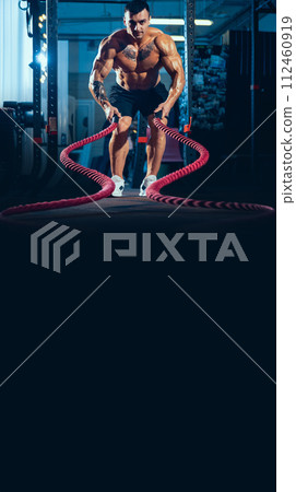 Young healthy man, athlete doing exercise with the ropes in the gym. Male model training hard his upper body Young healthy man, athlete doing exercise with the ropes in the gym. Male model training hard his upper body 112460919