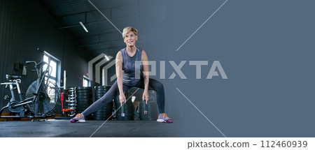 Smiling, positive, middle-aged woman training in gym, doing legs exercises, side lunges with dumbbells. Banner 112460939