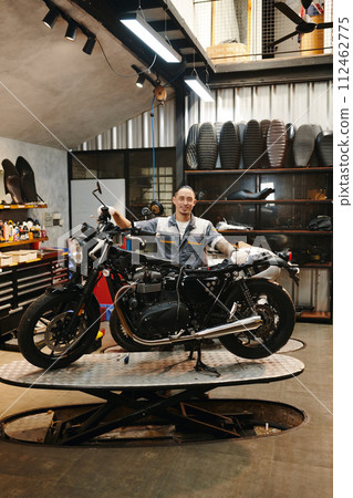 Proud smiling repairman standing next to motorcycle in his workshop Proud smiling repairman standing next to motorcycle in his workshop 112462775