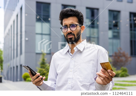 Close-up portrait of young upset Indian man businessman in white shirt standing near office building with chalk card and phone, worried spreading hands and looking at camera. 112464111