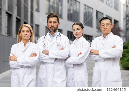 Portrait of an interracial group of doctors and scientists, men and women in white medical coats standing outside a clinic, crossing their arms over their chests and looking seriously at the camera. 112464135