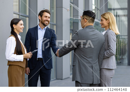 Business meeting of interracial businessmen. Men and women stand outside an office center and shake hands as a sign of getting to know each other and making a deal. Business meeting of interracial businessmen. Men and women stand outside an office center and shake hands as a sign of getting to know each other and making a deal. 112464148