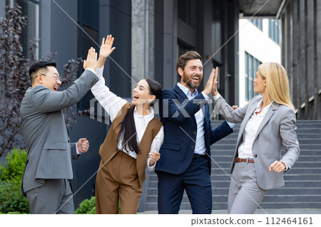 A young interracial business team is happy about a successful deal and partnership. A group of men and women stand outside an office center and high five each other, congratulating on victory. 112464161