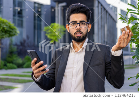 Portrait of a worried young Indian male businessman in a business suit sitting on a bench near the office, holding a credit card and a phone, looking disappointedly at the camera and spreading hands. Portrait of a worried young Indian male businessman in a business suit sitting on a bench near the office, holding a credit card and a phone, looking disappointedly at the camera and spreading hands. 112464213