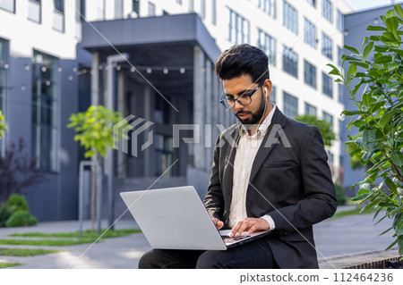 A young Indian man in a suit is sitting outside an office wearing headphones and a suit and is concentrating on his laptop. A young Indian man in a suit is sitting outside an office wearing headphones and a suit and is concentrating on his laptop. 112464236