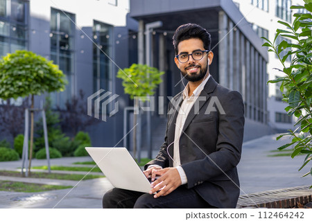 Portrait of a smiling young Indian man sitting in a business suit, wearing headphones, holding a laptop on his lap, looking confidently into the camera. 112464242