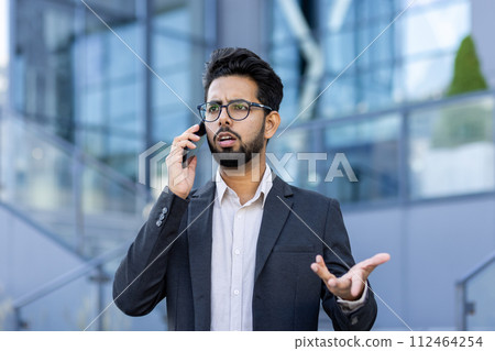 Close-up photo of an angry and sad young Indian man in a business suit standing on the street near an office center and talking ,emotionally on the phone while gesturing with his hands. Close-up photo of an angry and sad young Indian man in a business suit standing on the street near an office center and talking ,emotionally on the phone while gesturing with his hands. 112464254