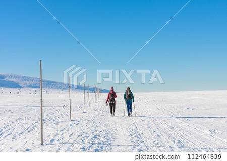 Two tourists walking in krkonose mountains, path from Silesian House to Meadow Hut, winter morning. Along the road are wooden long bars, tourist markings for winter season. 112464839