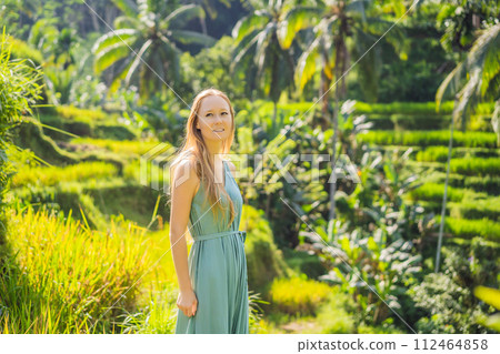 Beautiful young woman walk at typical Asian hillside with rice farming, mountain shape green cascade rice field terraces paddies. Ubud, Bali, Indonesia. Bali travel concept Beautiful young woman walk at typical Asian hillside with rice farming, mountain shape green cascade rice field terraces paddies. Ubud, Bali, Indonesia. Bali travel concept 112464858