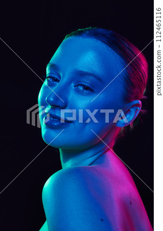 Close of young attractive woman looking at camera with smile in neon light against black studio background. Model with well-kept glowing skin. 112465116