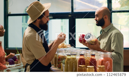 Storekeeper shows bulk products to man, presenting variety of pasta and spices to middle eastern customer. Young adult going grocery shopping at local zero waste supermarket. Handheld shot. 112465879