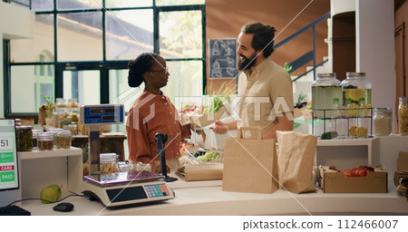 Vendor helping people with information about homegrown produce and sustainable lifestyle, presenting organic natural food choices. Couple looking for vegan alternatives at local eco store. Vendor helping people with information about homegrown produce and sustainable lifestyle, presenting organic natural food choices. Couple looking for vegan alternatives at local eco store. 112466007
