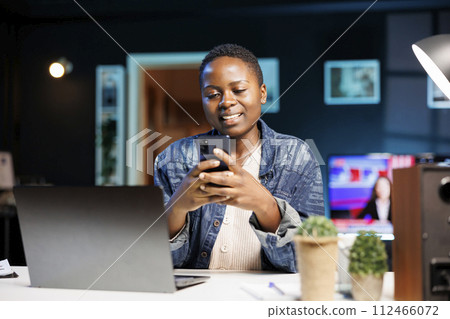 African american lady with positive emotion, using mobile device to surf the internet and chat with friends. Smiling black woman sitting in front of laptop, browsing social media on her smartphone. African american lady with positive emotion, using mobile device to surf the internet and chat with friends. Smiling black woman sitting in front of laptop, browsing social media on her smartphone. 112466072