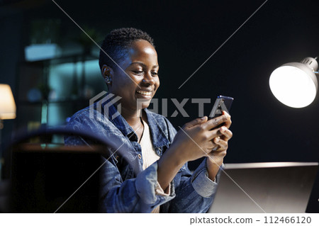 Cheerful black woman utilizing modern technology, taking break from laptop and looking at social media on mobile device. African american lady surfing the net on cellphone, online communication. Cheerful black woman utilizing modern technology, taking break from laptop and looking at social media on mobile device. African american lady surfing the net on cellphone, online communication. 112466120