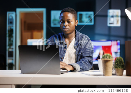 Busy black female freelancer sitting in her modern living room office, working on personal computer. African american college student typing and reviewing information on digital laptop. Busy black female freelancer sitting in her modern living room office, working on personal computer. African american college student typing and reviewing information on digital laptop. 112466138