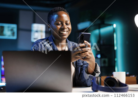 Cheerful female college student using mobile device to engage with friends on social media platforms. African american lady using cell phone to surf the web while sitting at desk in her home office. Cheerful female college student using mobile device to engage with friends on social media platforms. African american lady using cell phone to surf the web while sitting at desk in her home office. 112466153