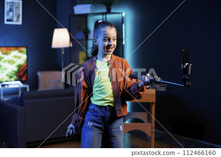 Generation Z media star with cellphone on selfie stick, filming herself doing room tour for children watching her online content. Kid doing video presenting her home to social media channels audience Generation Z media star with cellphone on selfie stick, filming herself doing room tour for children watching her online content. Kid doing video presenting her home to social media channels audience 112466160