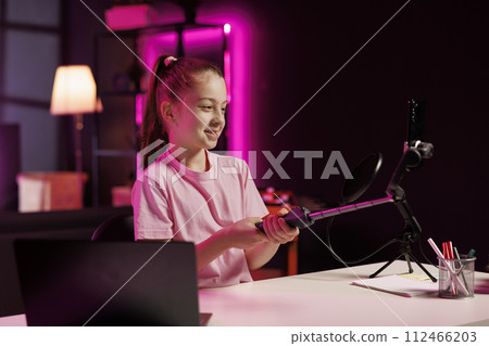 Cute girl filming room tour inside home studio using smartphone on selfie stick. Little kid doing video presenting her neon lit apartment to interested children audiences Cute girl filming room tour inside home studio using smartphone on selfie stick. Little kid doing video presenting her neon lit apartment to interested children audiences 112466203