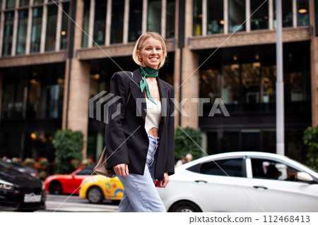 Beautiful young stylish woman walking down city street against backdrop of building, wearing fashionable clothes jeans, black blazer, handbag. Happy Fashion model Beautiful young stylish woman walking down city street against backdrop of building, wearing fashionable clothes jeans, black blazer, handbag. Happy Fashion model 112468413