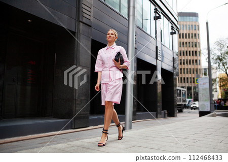 Stylish fashion woman in a pink suit, shirt and shorts, wearing heels, walking confidently against a city backdrop. Urban street style allure 112468433