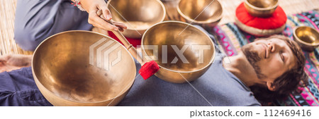 BANNER, LONG FORMAT Nepal Buddha copper singing bowl at spa salon. Young beautiful man doing massage therapy singing bowls in the Spa against a waterfall. Sound therapy, recreation, meditation 112469416