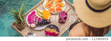 BANNER, LONG FORMAT Breakfast tray in swimming pool, floating breakfast in luxury hotel. Girl relaxing in the pool drinking smoothies and eating fruit plate, smoothie bowl by the hotel pool. Exotic 112469454