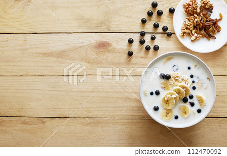 Oatmeal porridgewith bananas, nuts, raisins, blueberries and milk on table on wooden background. Healthy breakfast and diet food. Oatmeal porridgewith bananas, nuts, raisins, blueberries and milk on table on wooden background. Healthy breakfast and diet food. 112470092